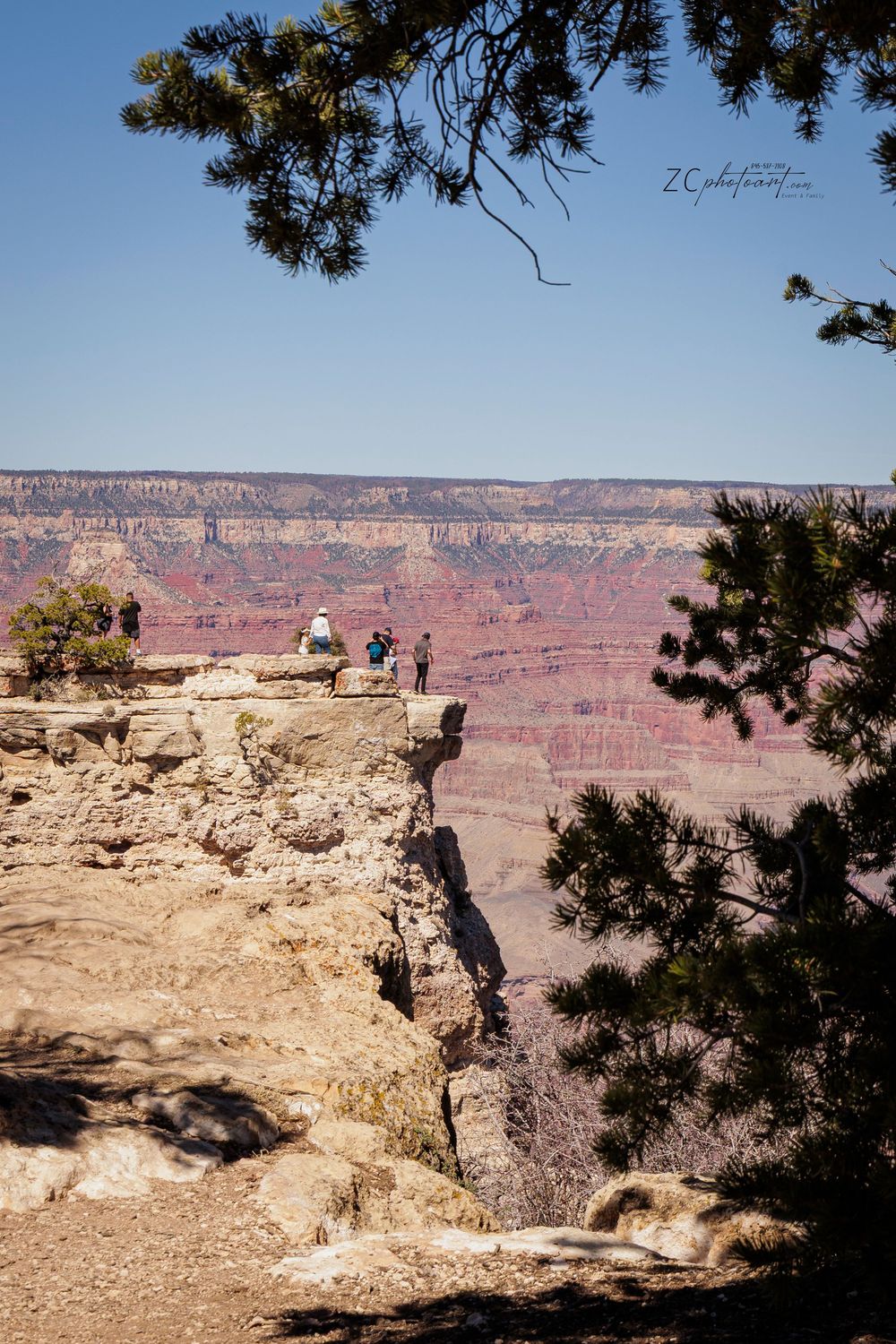Tourists stand on rocky cliff edge overlooking the vast, colorful Grand Canyon landscape under a clear blue sky.