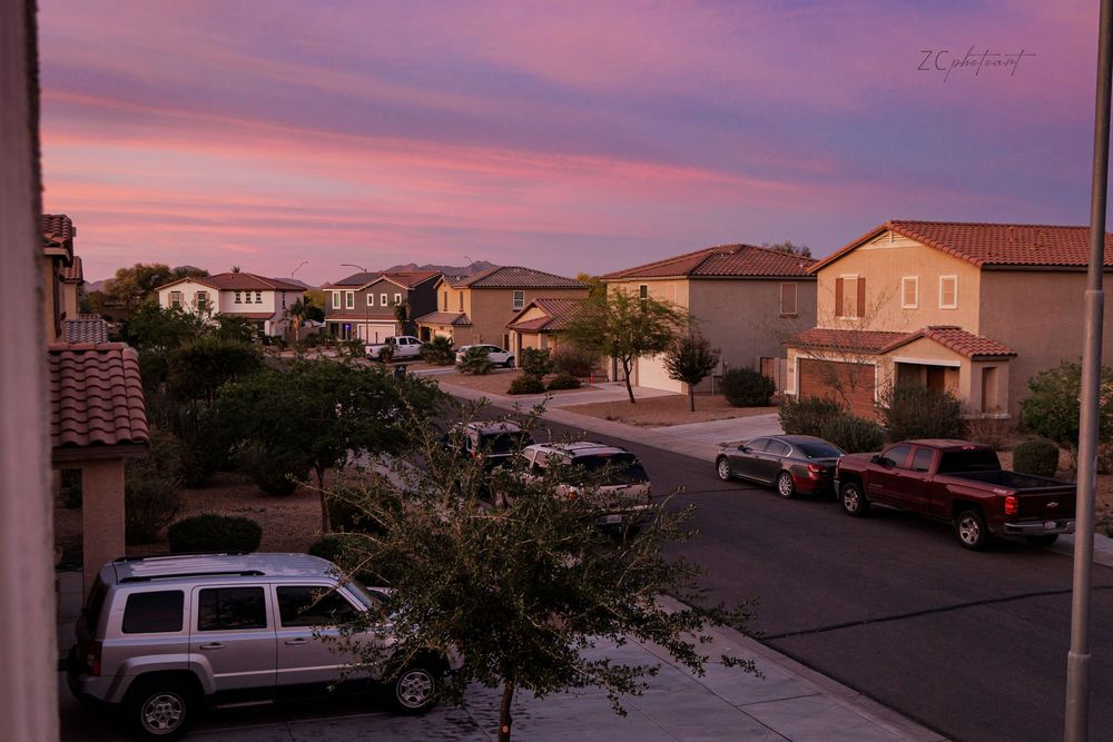 Suburban neighborhood street at sunset with pink and purple sky, parked cars, and tan stucco homes in a desert community.