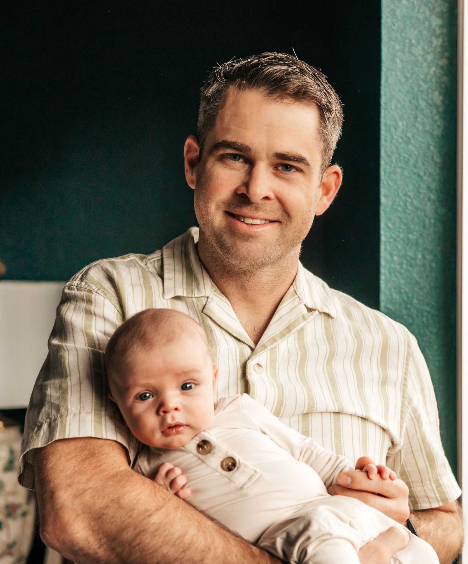 A smiling man holds a young baby dressed in white, posing together in a warmly lit room  | Newborn Photographer in Broomfield Colorado