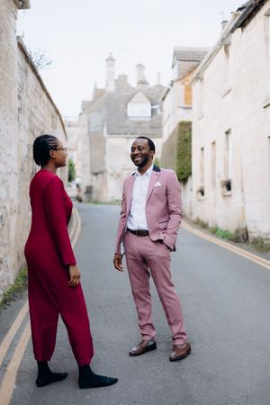 Stylish Black couple posing on a quiet street, man in pink suit, woman in red jumpsuit in Painswick near Stroud.