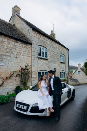 Bride and groom posing beside a white Audi R8 in front of a stone cottage in the Cotswolds for their engagement photoshoot.