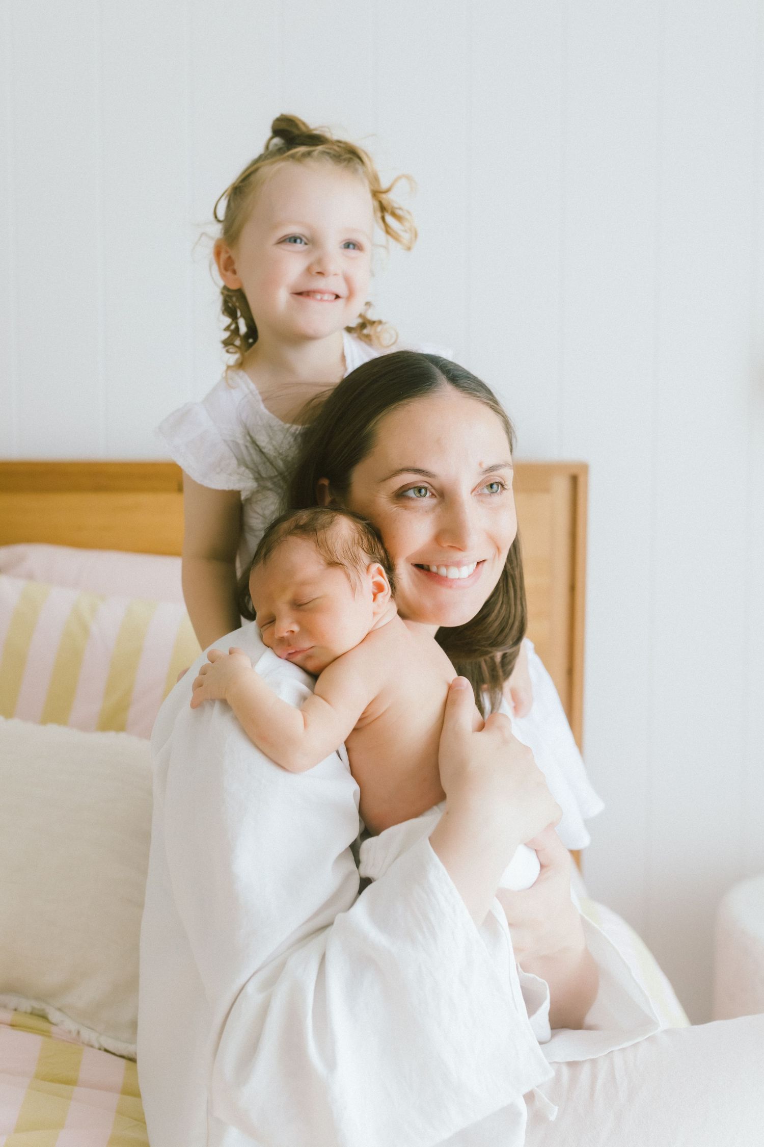 A smiling mother in white holds a sleeping newborn while her curly-haired toddler daughter perches behind her on a bed.