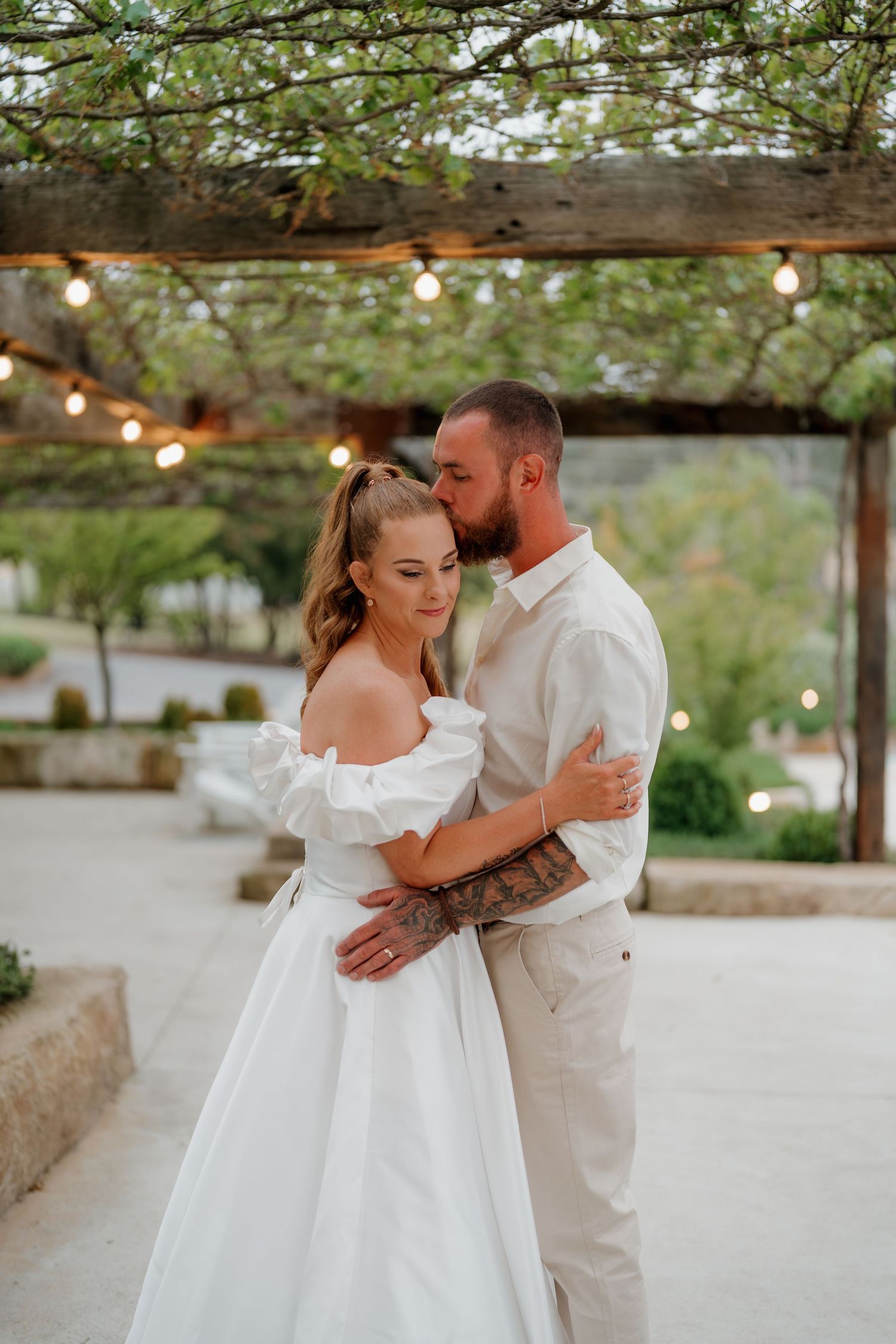 Bride and groom share a romantic embrace under a pergola with string lights at an outdoor wedding venue.