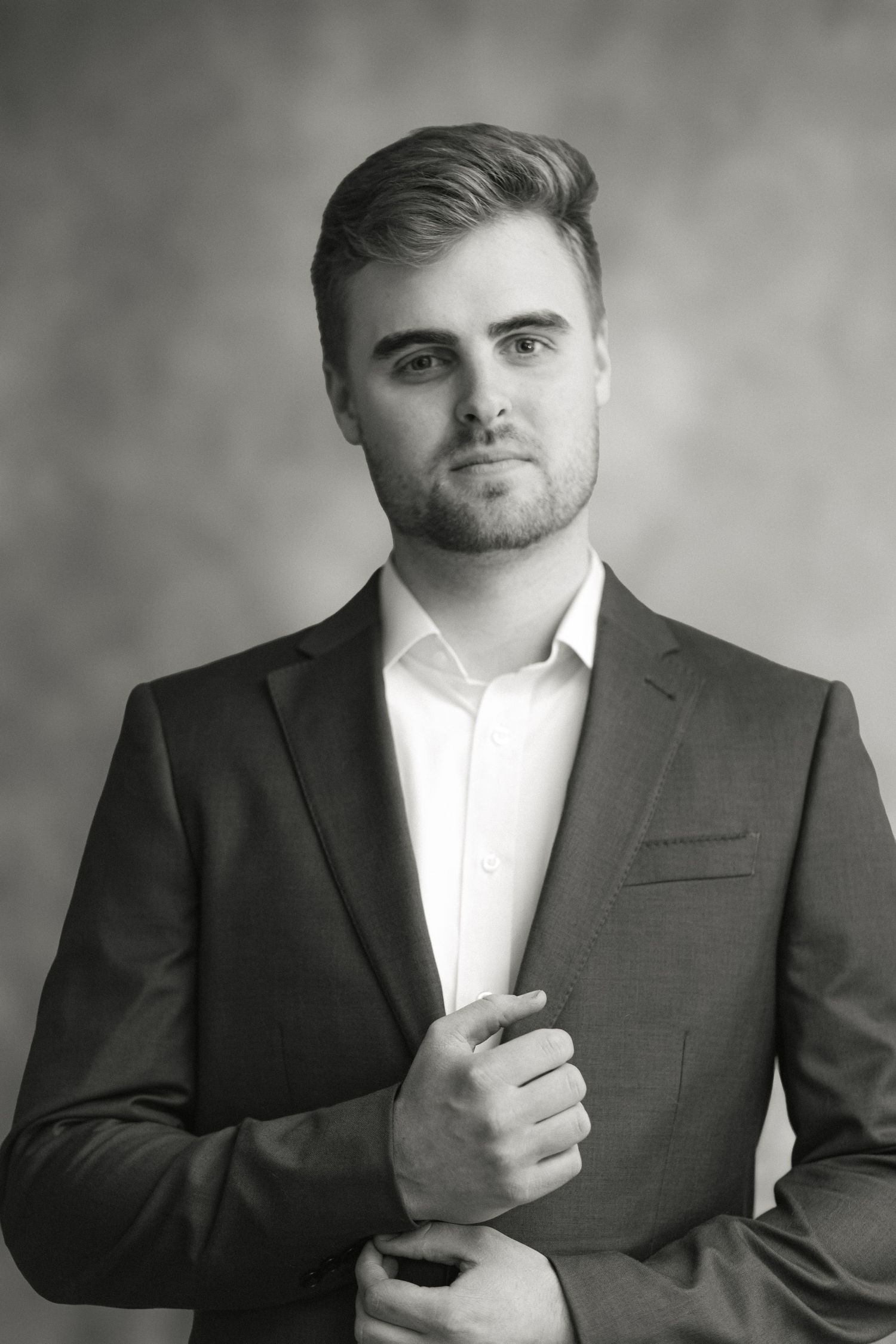 Young professional man in dark suit adjusts jacket sleeve in black and white studio portrait.