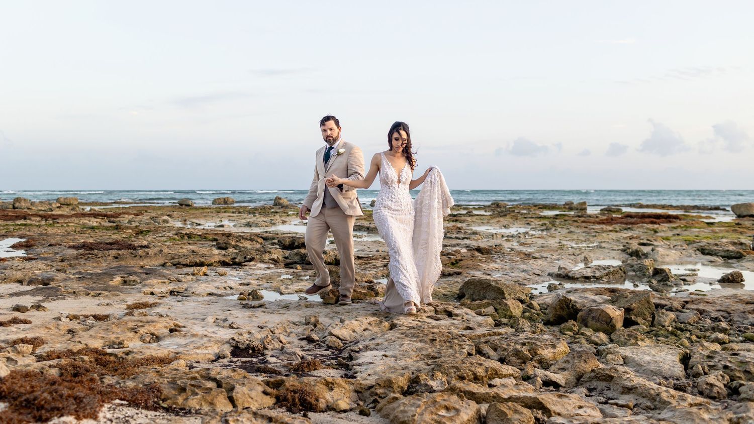 Couple in wedding attire walks hand-in-hand on rocky beach shoreline with ocean backdrop at dusk.