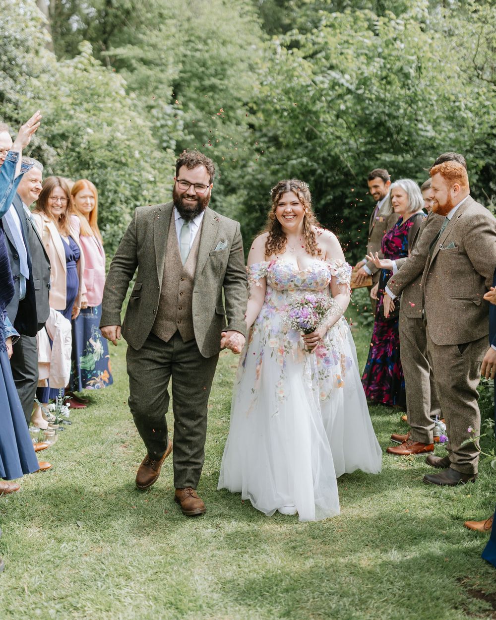 A joyful couple walks hand-in-hand at an outdoor wedding ceremony, surrounded by cheering guests in a lush garden.