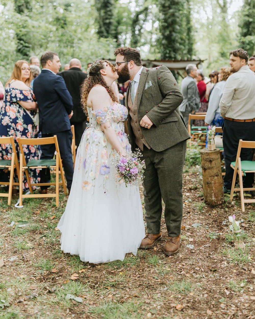 Newlywed couple sharing a first kiss in an outdoor woodland wedding ceremony with guests seated on wooden chairs.