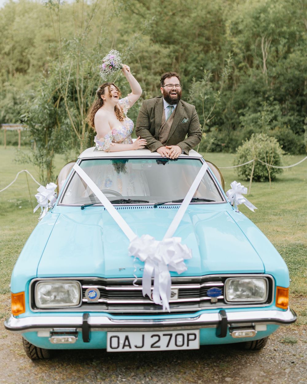Happy newlyweds celebrating in a vintage turquoise Ford car decorated with white ribbons on their wedding day.