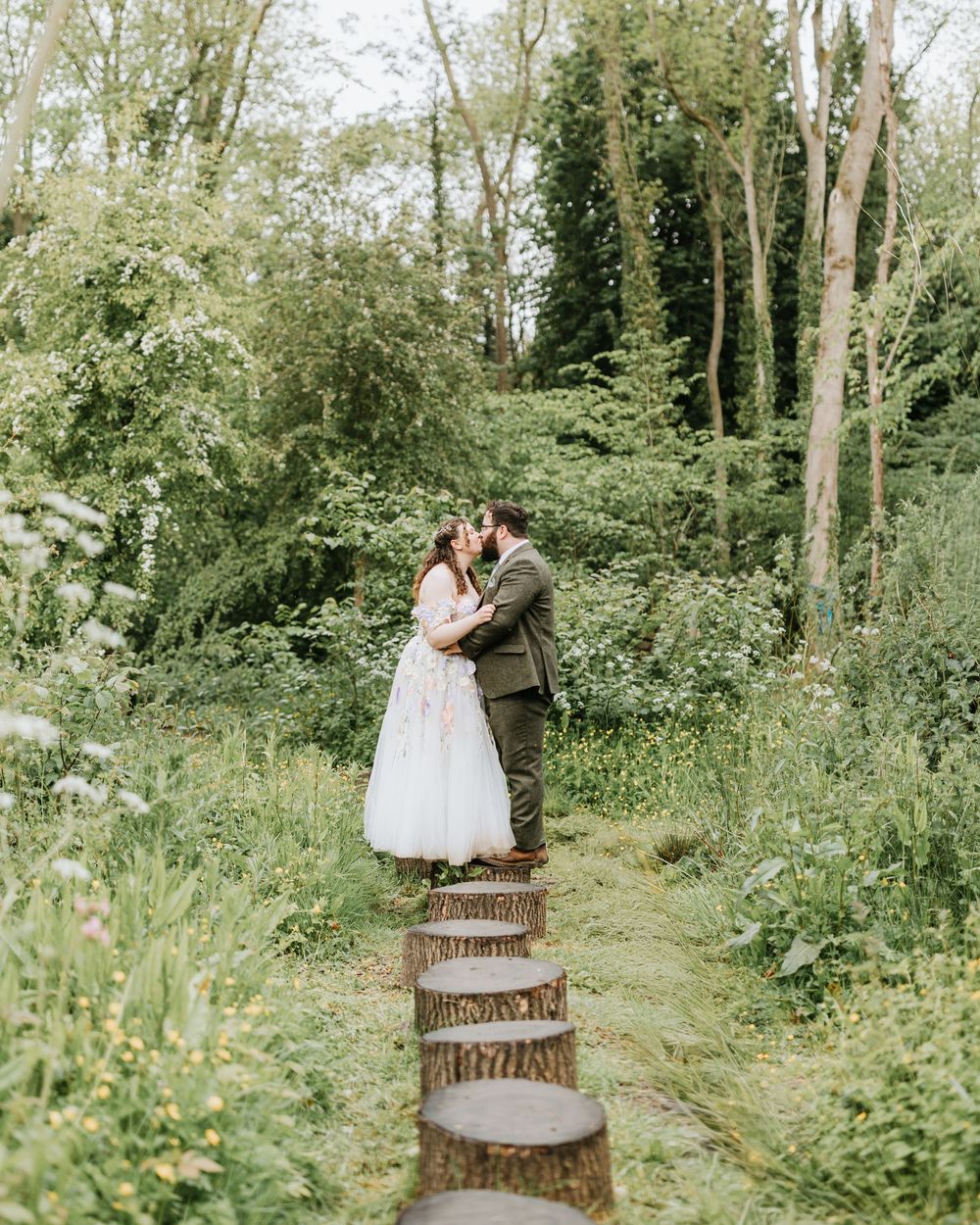 A couple shares a romantic kiss in a lush green woodland garden, with tree stump stepping stones leading to them.
