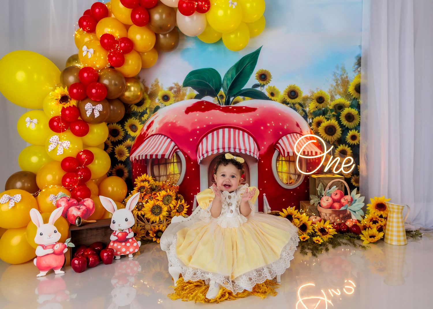Baby girl in yellow dress sits before a Snow White apple house backdrop with balloons and sunflowers at a first birthday cake smash.