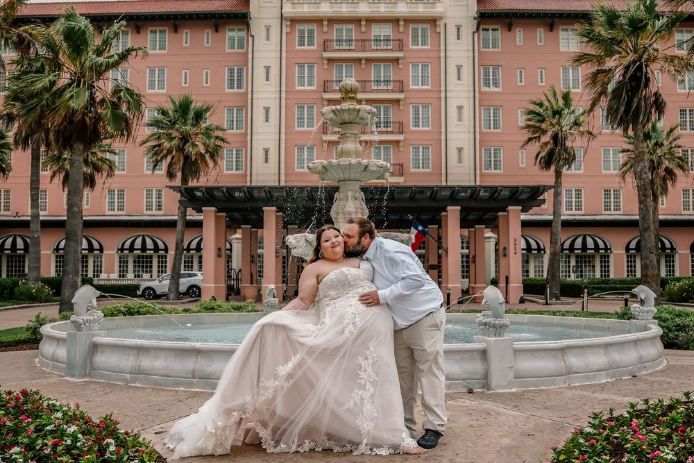 Bride and groom share a romantic kiss near a fountain outside a grand pink hotel surrounded by palm trees.