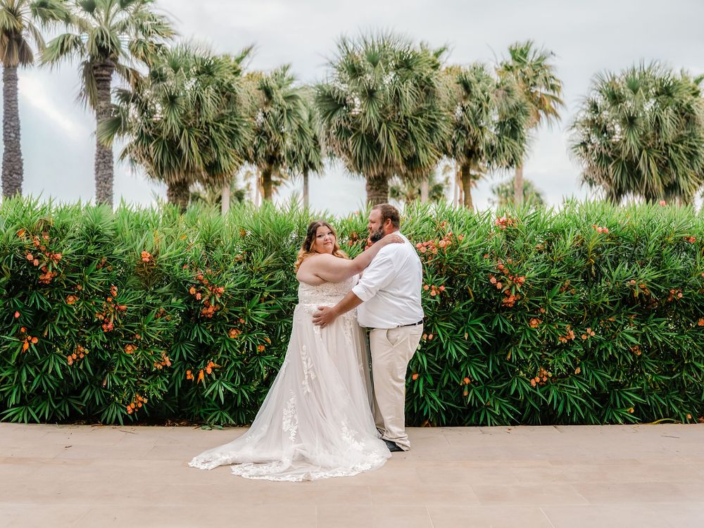 Bride and groom embrace outdoors in front of a lush tropical hedge with colorful flowers and palm trees.