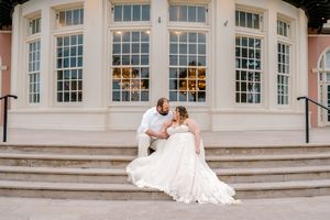 Bride in a flowing gown sits with groom on steps outside an elegant white building with large windows.