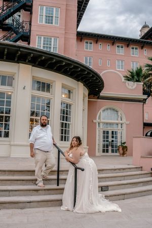 Bride and groom pose on steps outside a historic pink building with white trim and arched windows.