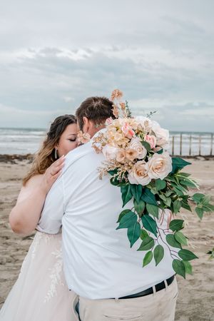 Bride and groom embrace on beach holding a blush floral bouquet with roses and greenery, ocean waves in background.