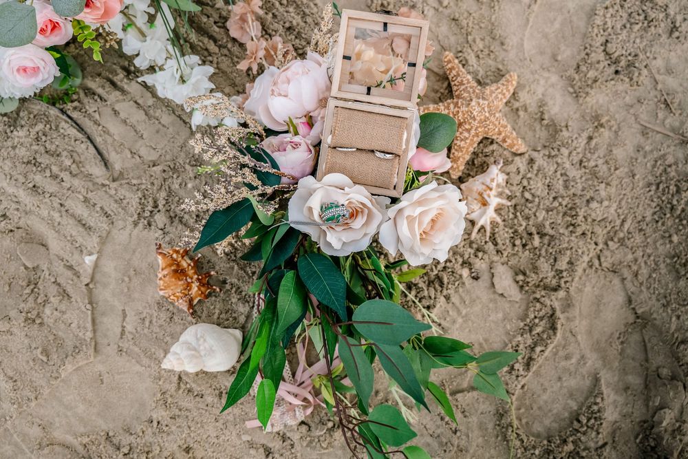 Beach wedding flat lay with blush roses, peonies, greenery, starfish, seashells, and ring box on sand.