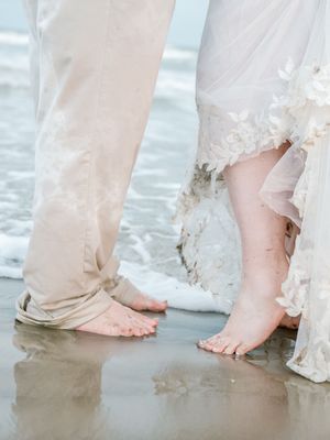 Barefoot bride and groom standing at ocean shoreline, wedding dress hem and khaki pants getting wet in the surf.