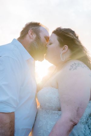 Bride and groom sharing a romantic kiss backlit by golden sunset light, bride wearing lace strapless gown with hoop earrings.