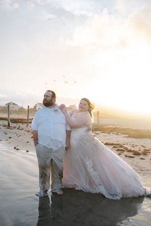 Bride in blush ballgown and groom in white shirt pose on wet beach at golden sunset with seagulls overhead.