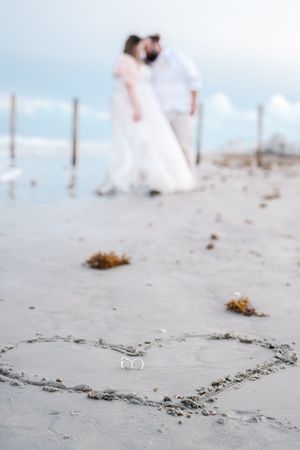 Heart drawn in beach sand with blurred bride and groom kissing in background near wooden posts at the ocean.