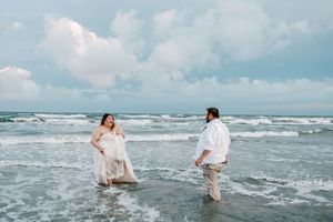 Bride and groom wading through ocean waves on the beach, bride in flowing white gown smiling at groom.