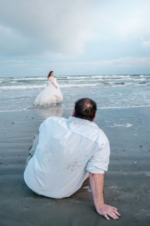 Groom sitting on wet sand watching bride in white wedding gown standing in ocean waves on the beach.