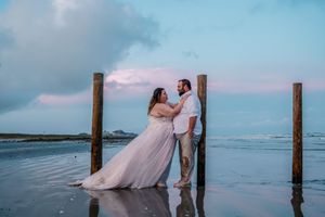 Bride in blush gown and groom lean against wooden pier posts on reflective beach at pink and blue sunset.