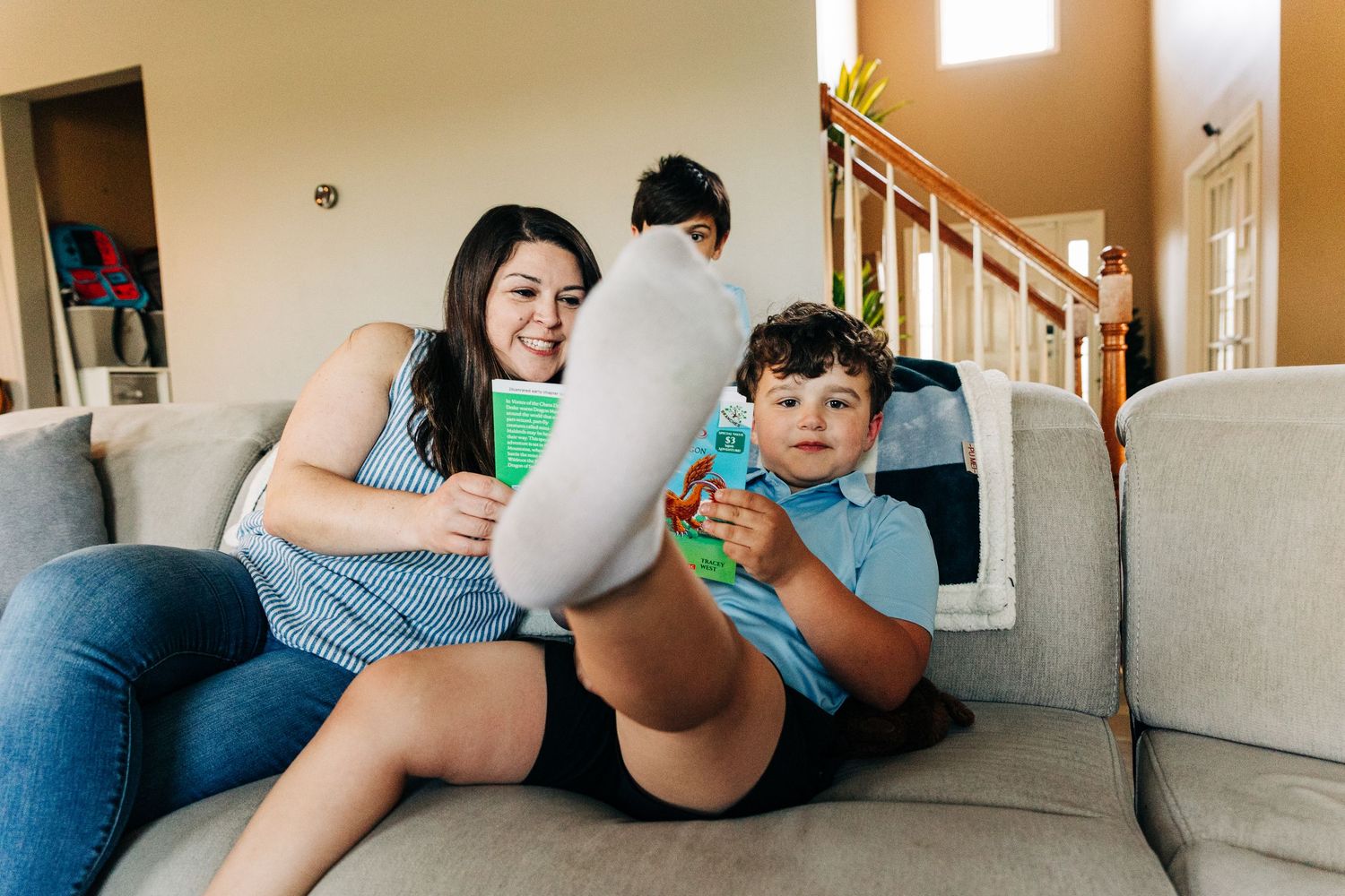 A woman and young boy sit on a couch together, smiling and holding books in a cozy home living room.