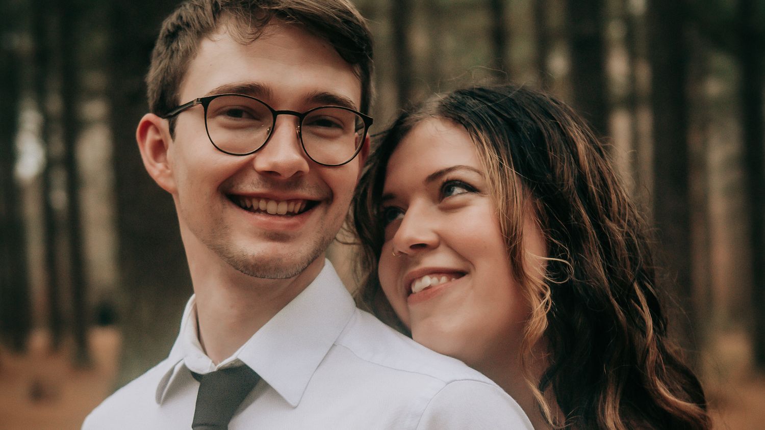 Smiling young couple posing together in a pine forest, man wearing glasses and tie.