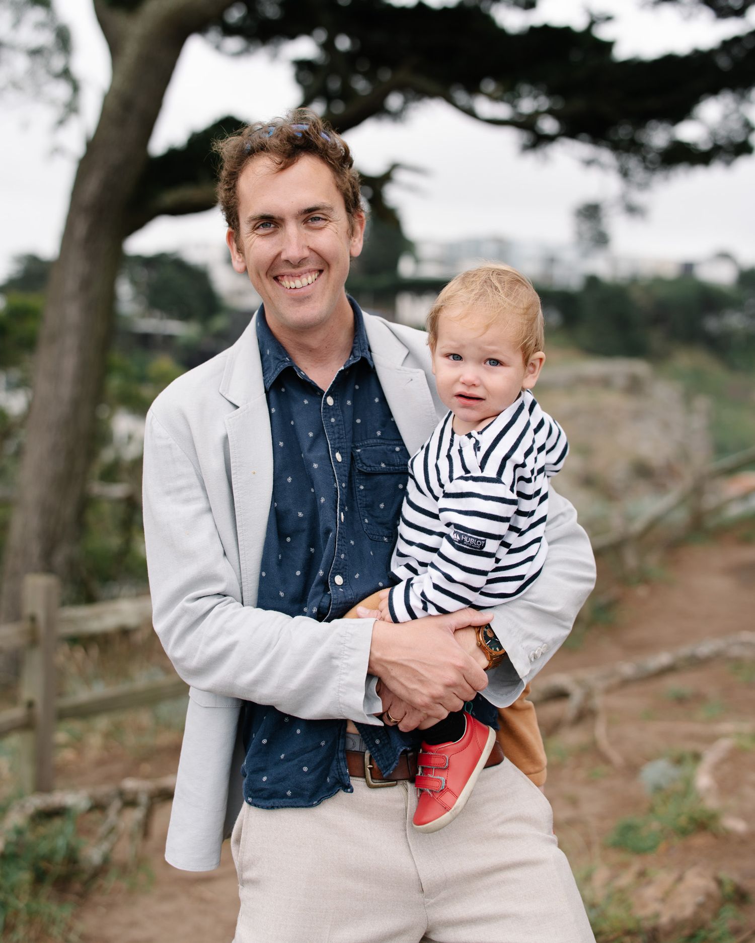 A man smiling while holding a young girl in a red striped shirt near the beach.