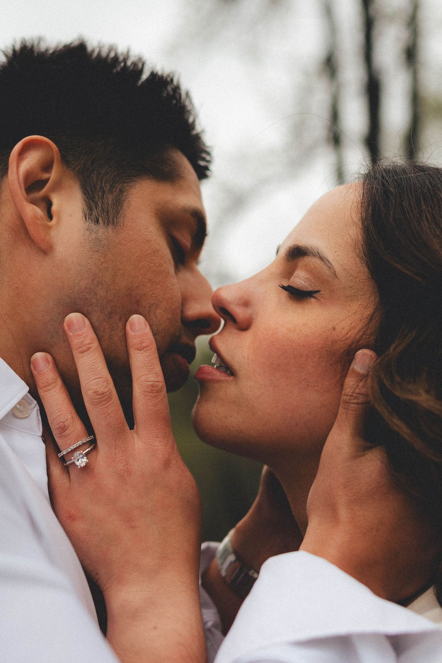 Couple sharing a romantic kiss outdoors, woman wearing diamond engagement ring, both dressed in white shirts.