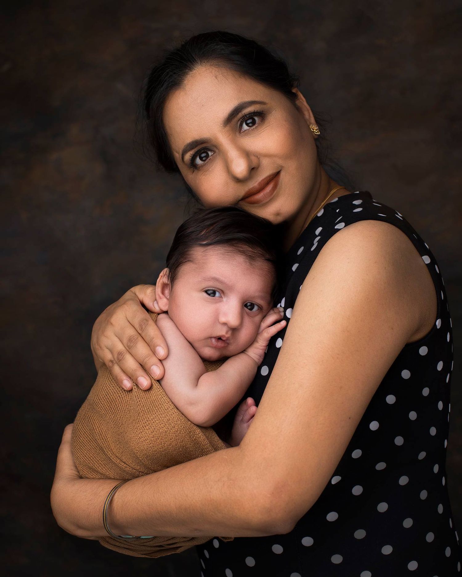 A mother in a polka dot dress tenderly holds her swaddled newborn baby in a warm studio portrait.
