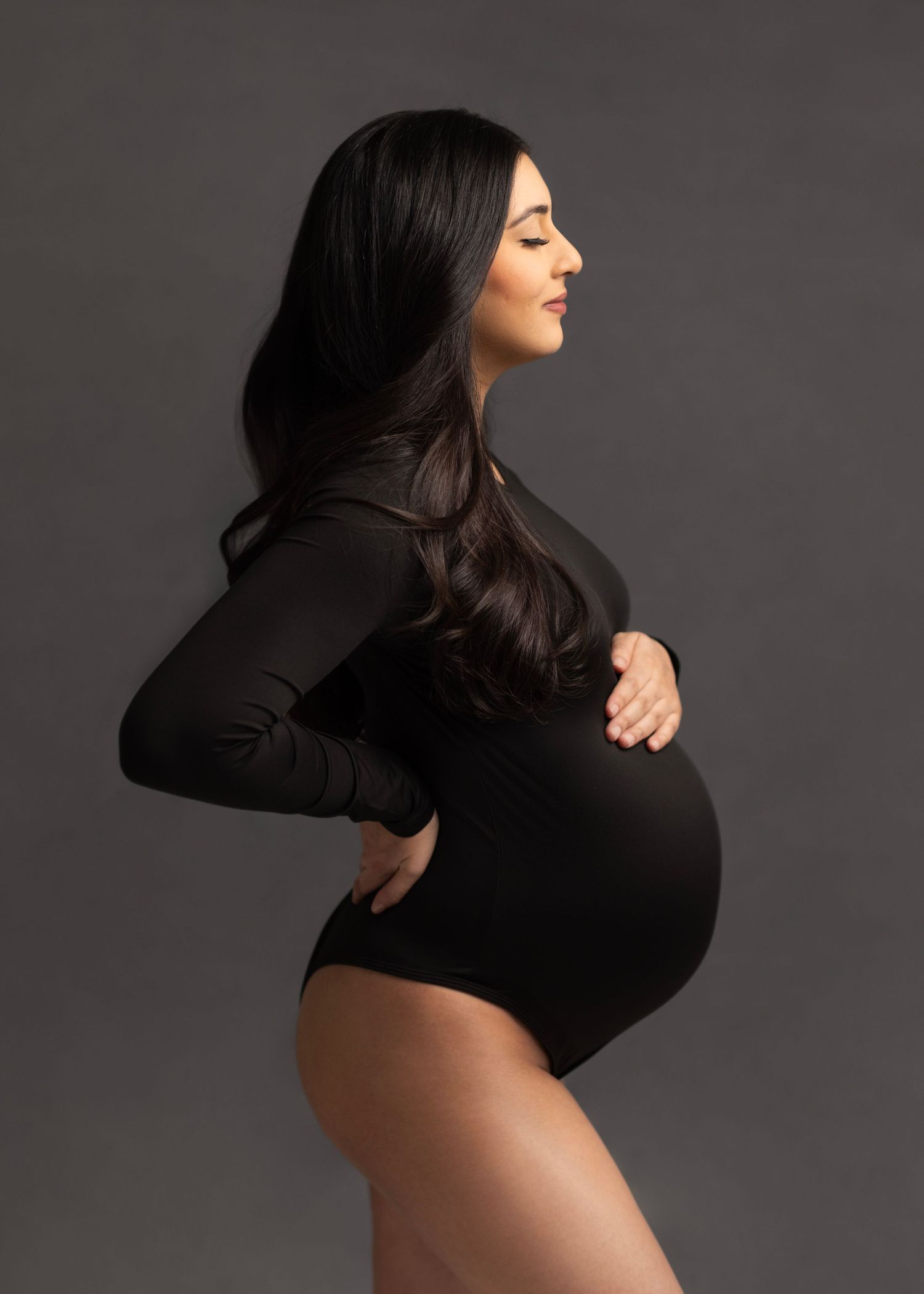 Pregnant woman in black bodysuit posing sideways, cradling baby bump in professional maternity studio photo.