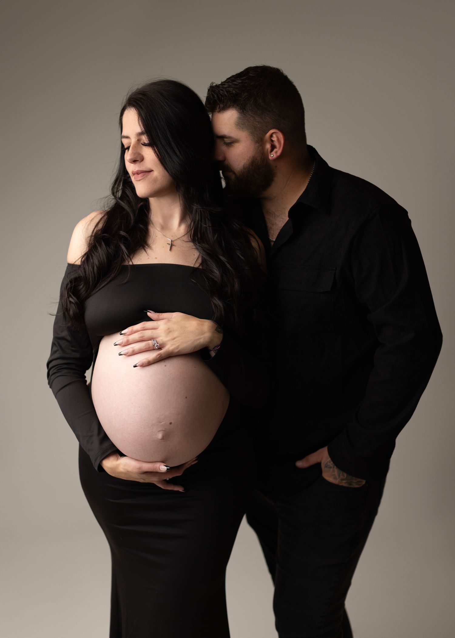 Couple in black outfits posing for maternity photo, man kissing woman's head while she cradles her baby bump in studio.