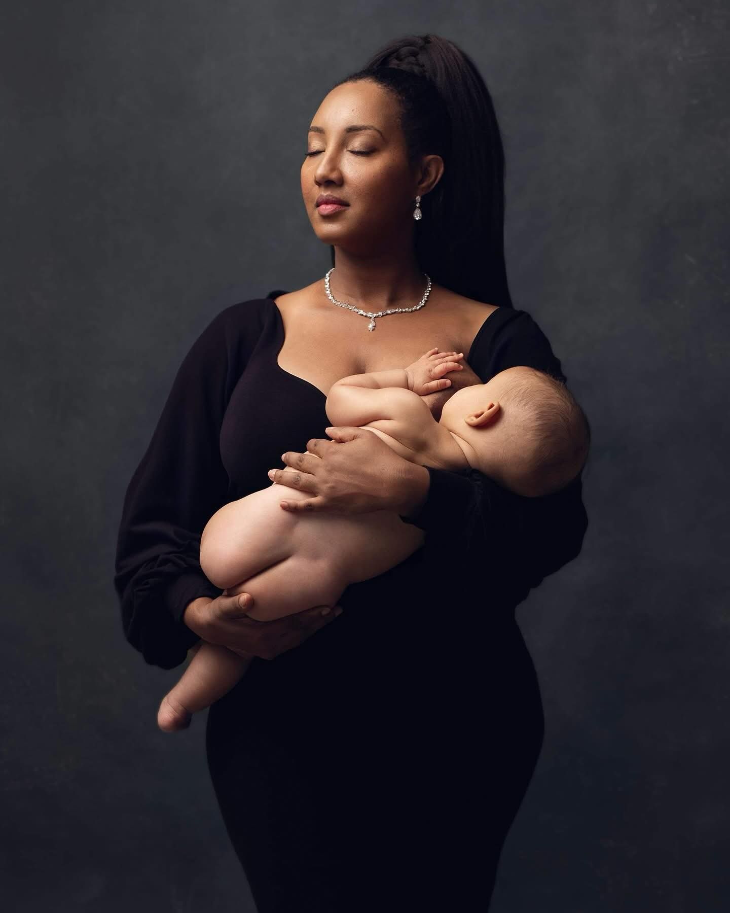 Elegant woman in black dress holding a newborn baby, wearing pearl jewelry against a dark studio background.