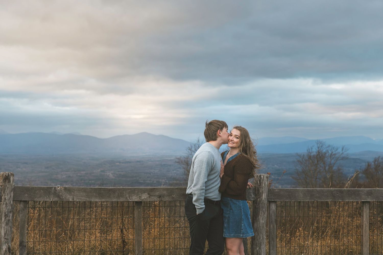 Couple sharing a romantic kiss at a scenic mountain overlook with dramatic cloudy skies and valley views.