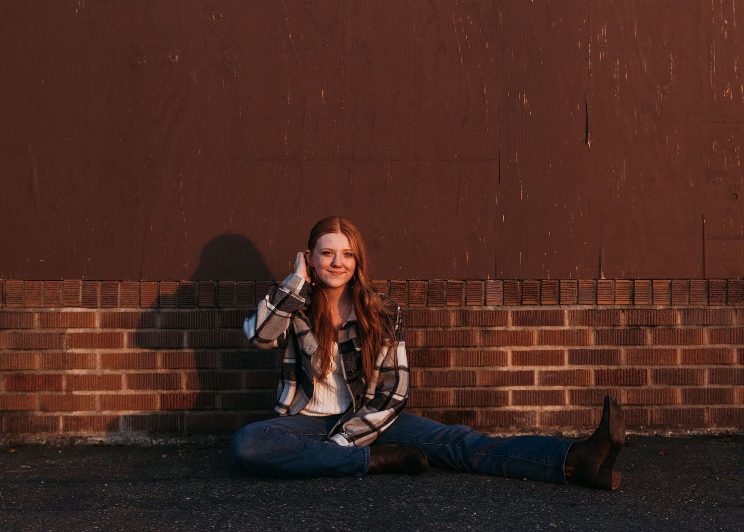 Young woman with red hair sits against a brick wall outdoors, wearing a plaid shirt and jeans, smiling warmly.