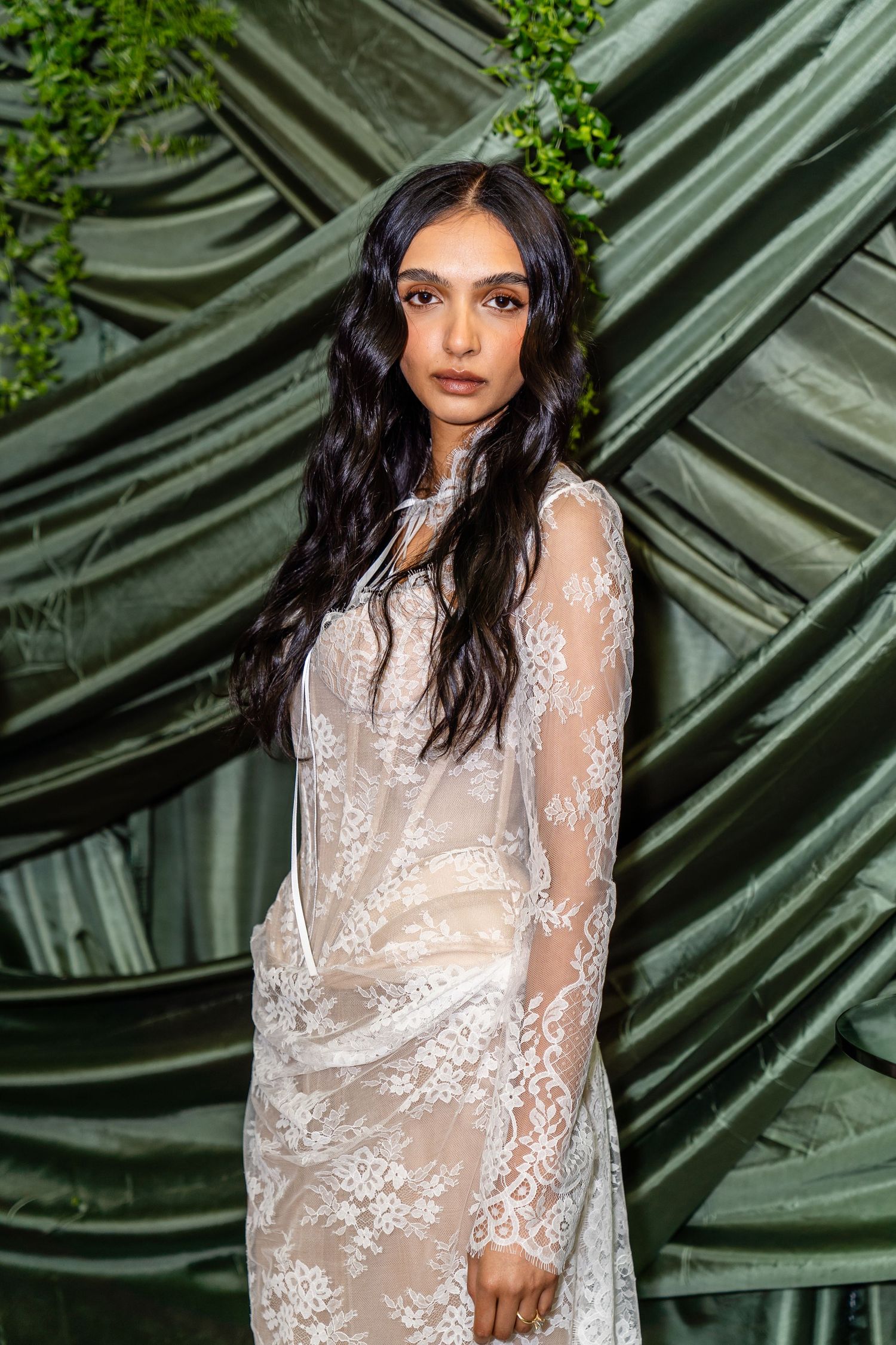 Woman in sheer white lace gown posing against draped green fabric backdrop with foliage.