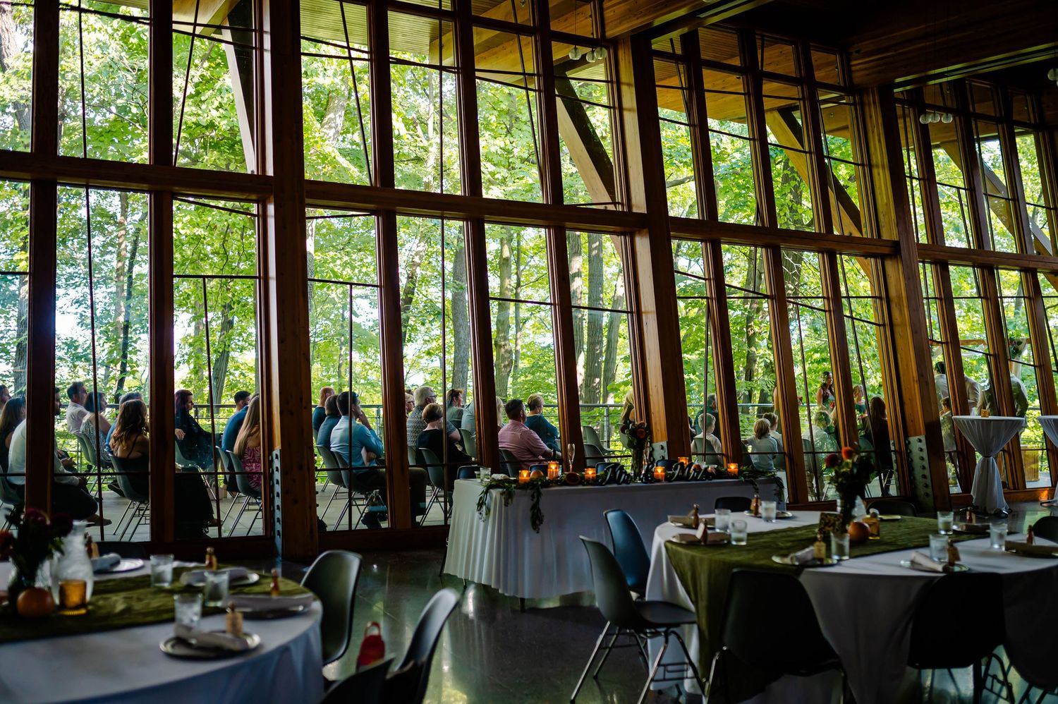 Elegant event venue with floor-to-ceiling windows overlooking lush forest, guests dining at tables with green linens.