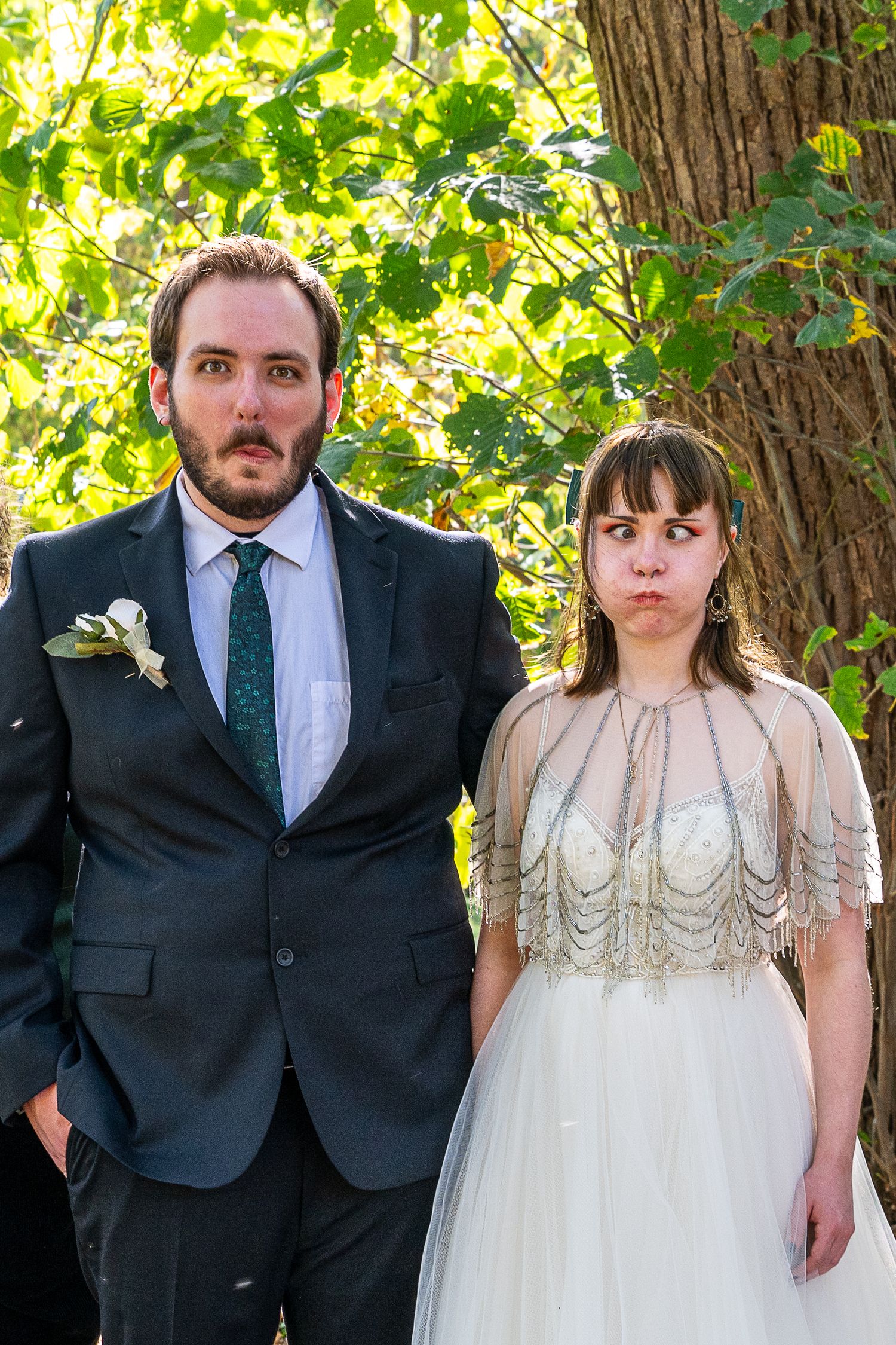 Bride and groom making funny faces outdoors near a tree, wearing wedding attire including a beaded bridal cape.
