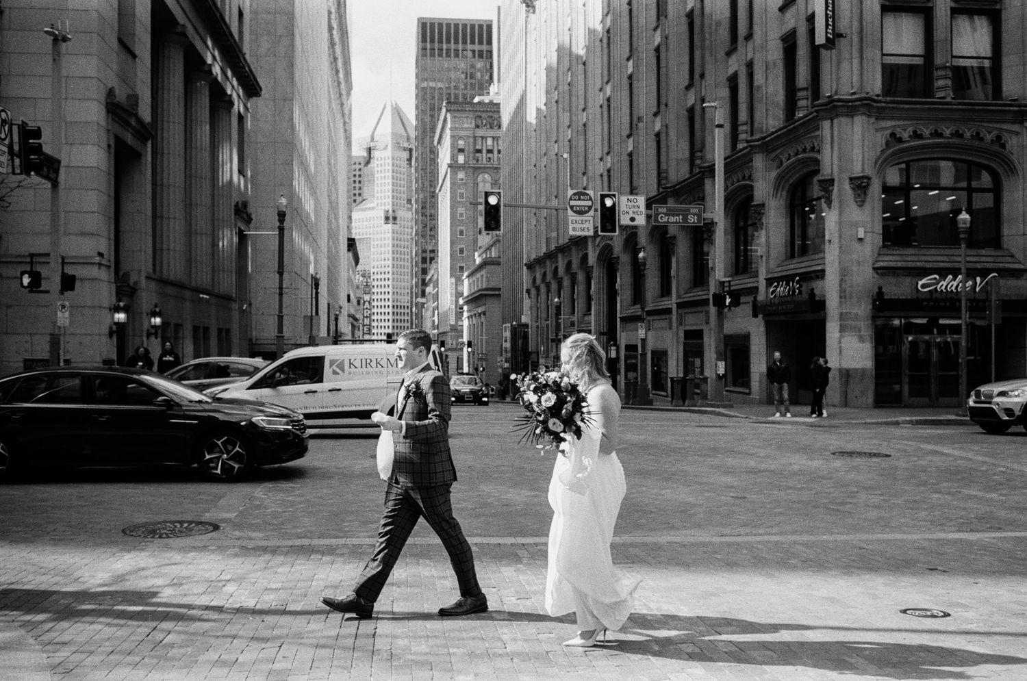 Black and white photo of a bride and groom crossing a city street, bride holding a bouquet.