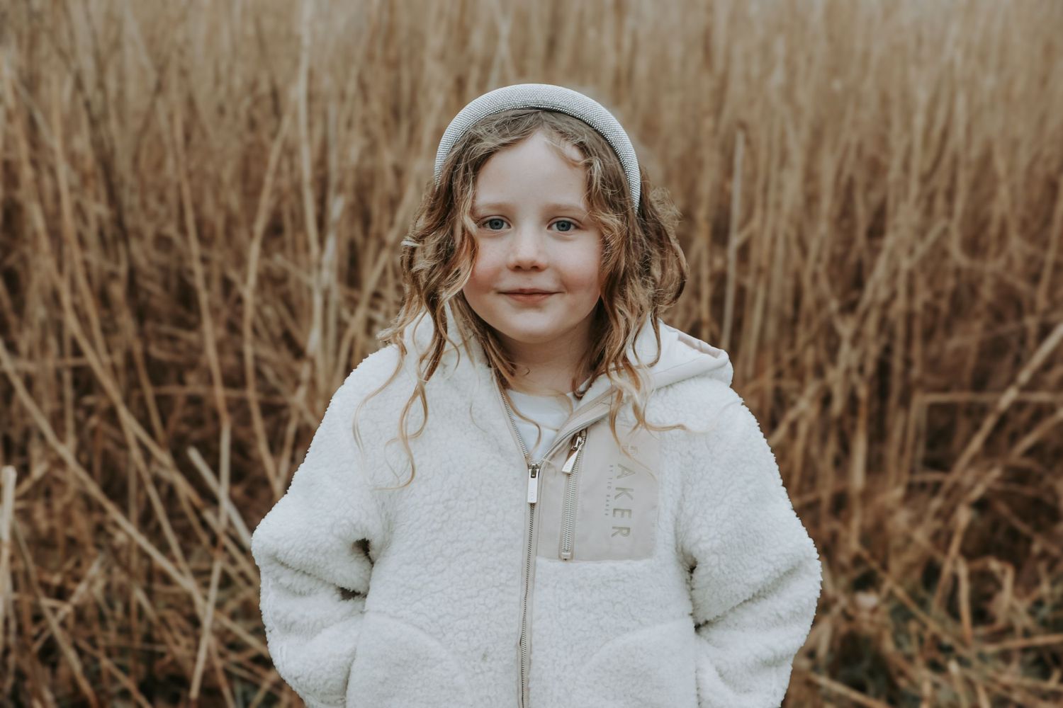 Young person in white fuzzy jacket standing in tall brown grass on an overcast day.