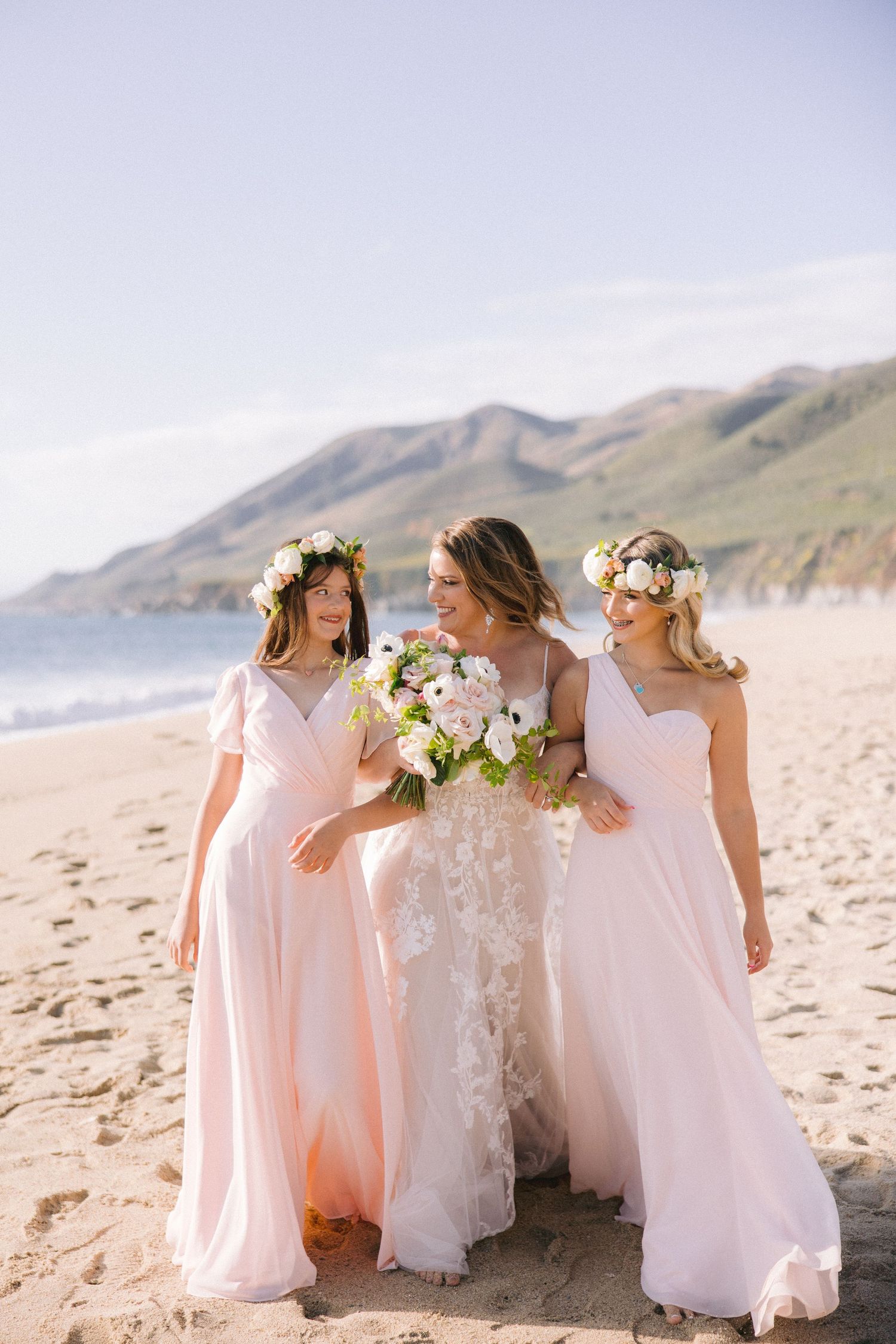 Three women in flowing pink and white dresses walk together on a scenic beach with mountains in the background.