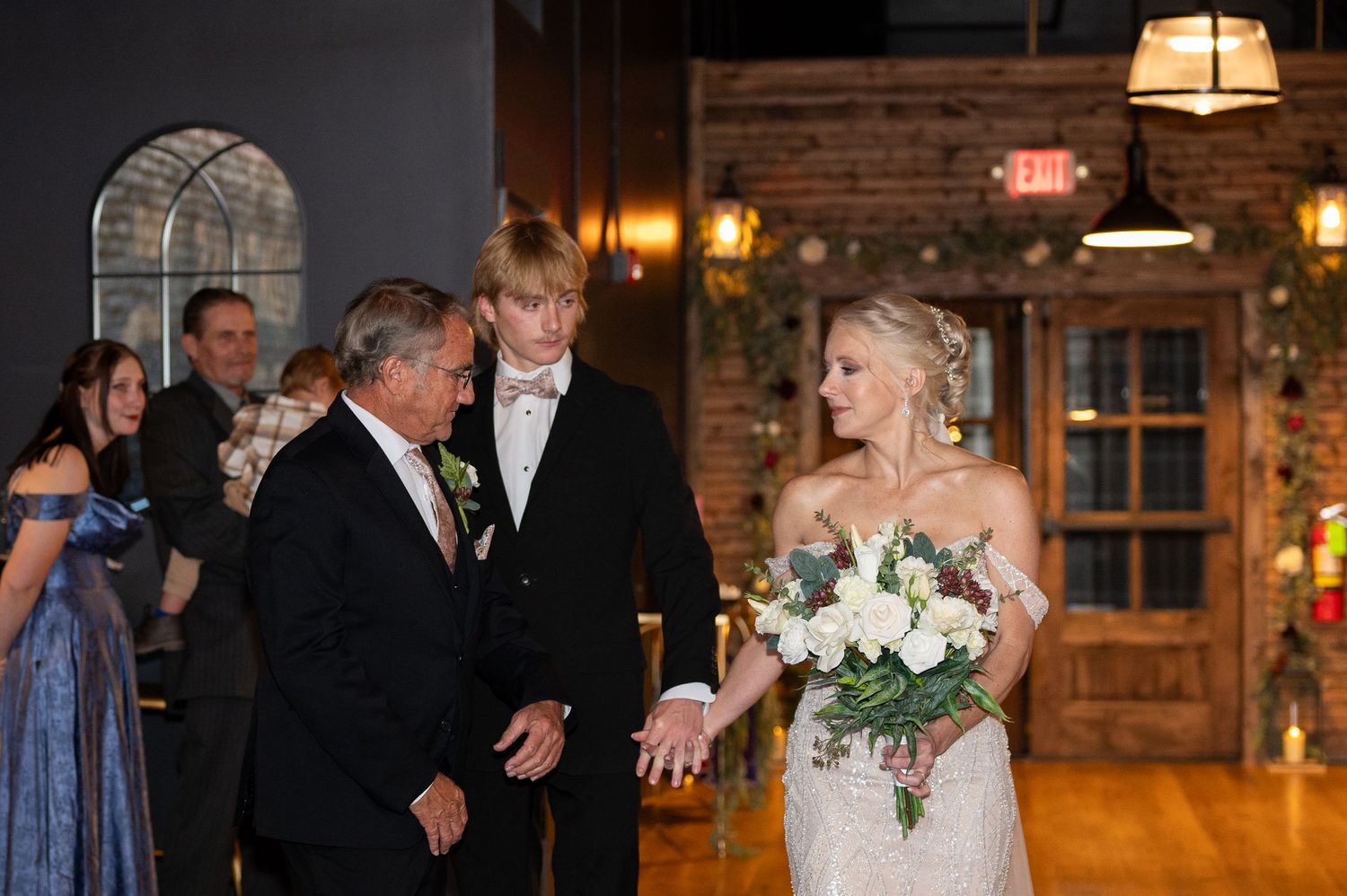 A heartfelt moment during a rustic evening wedding ceremony with white floral bouquet in a warmly lit wooden venue.