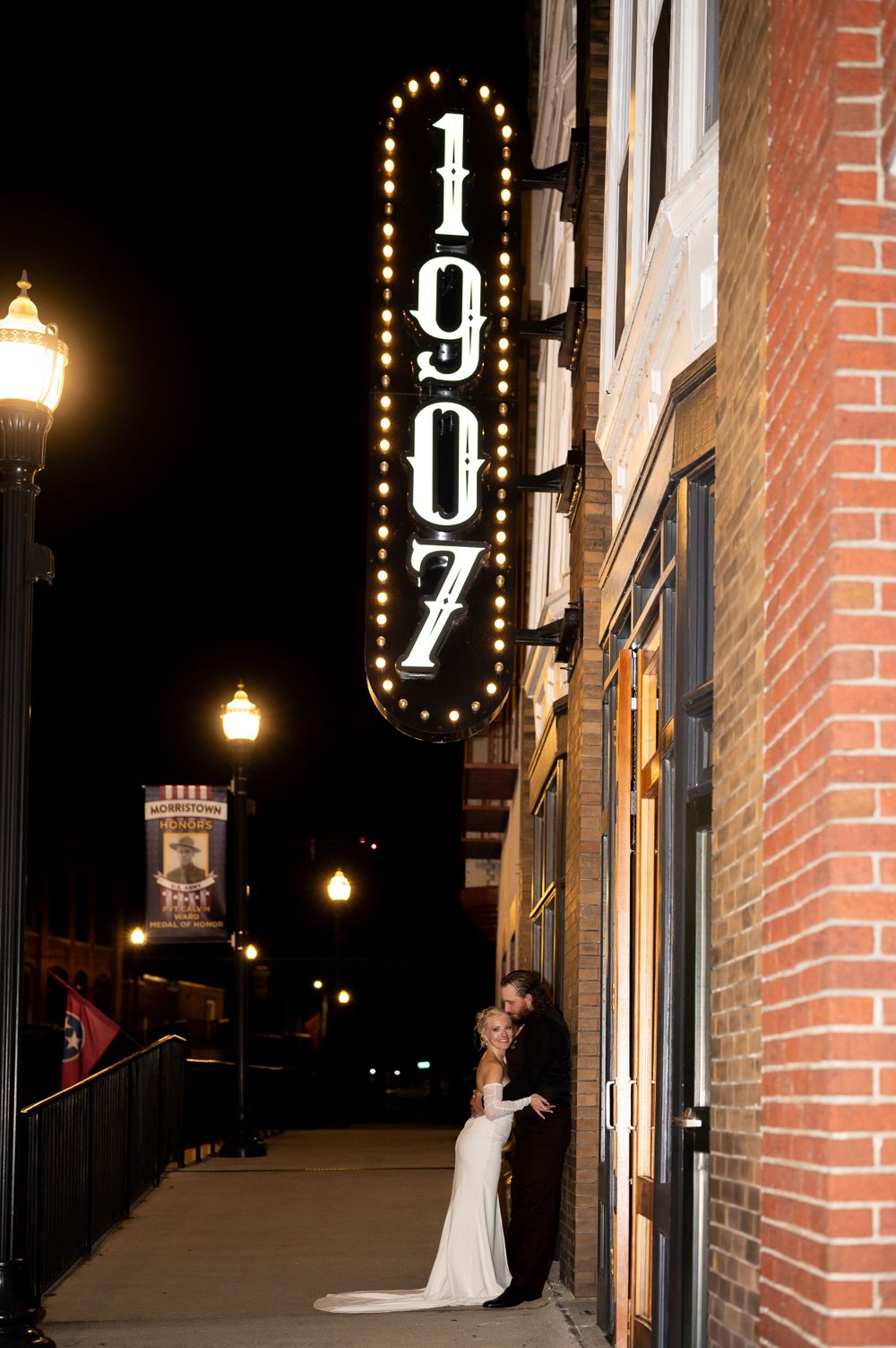 A lit vertical 1907 sign glows against a brick building at night with illuminated street lamps along a sidewalk.