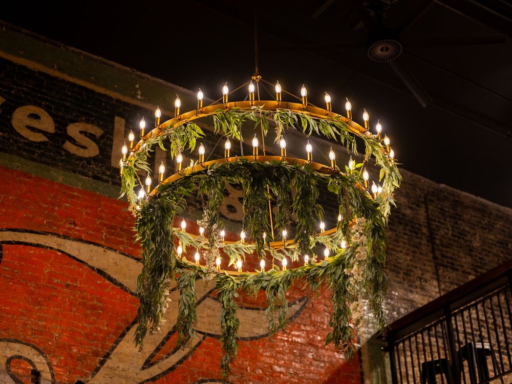 Ornate antique chandelier with glowing bulbs hangs against exposed brick wall in rustic industrial setting.
