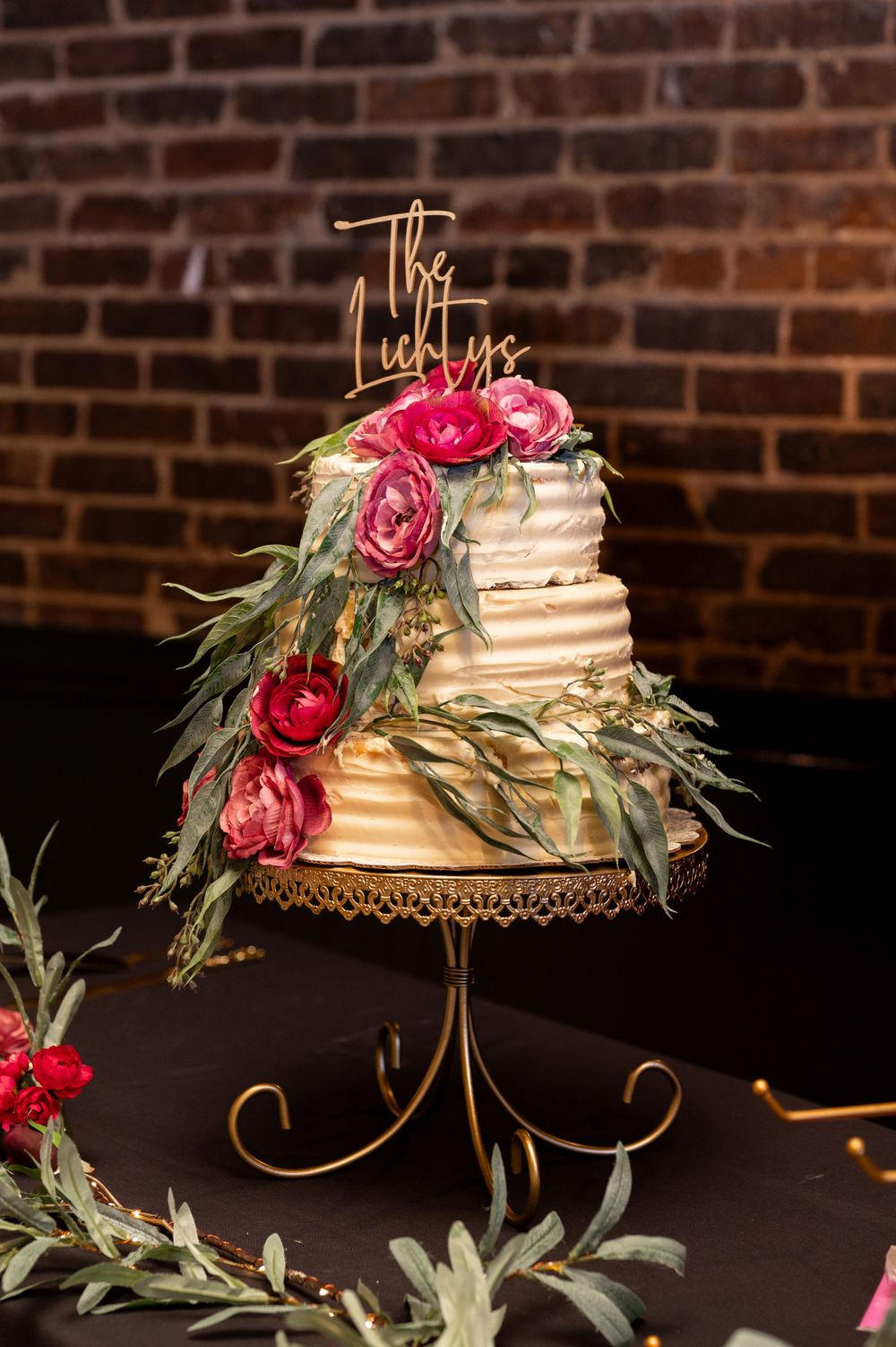 Elegant white wedding cake decorated with pink roses and eucalyptus leaves on a gold metal stand against brick wall.