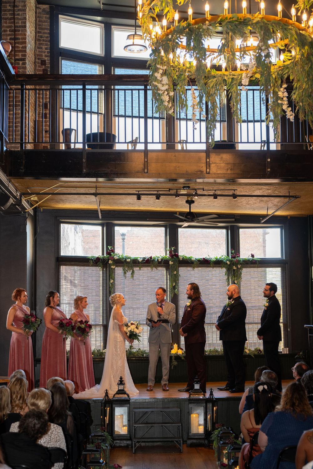 Indoor wedding ceremony takes place on elevated platform under rustic chandelier with lush greenery in industrial venue space.