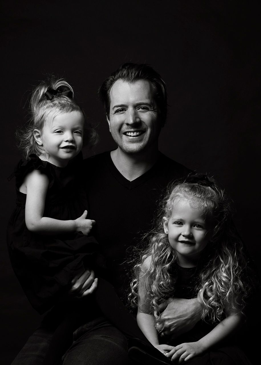 A black and white studio portrait of a smiling parent with two young children in casual attire against a dark background.