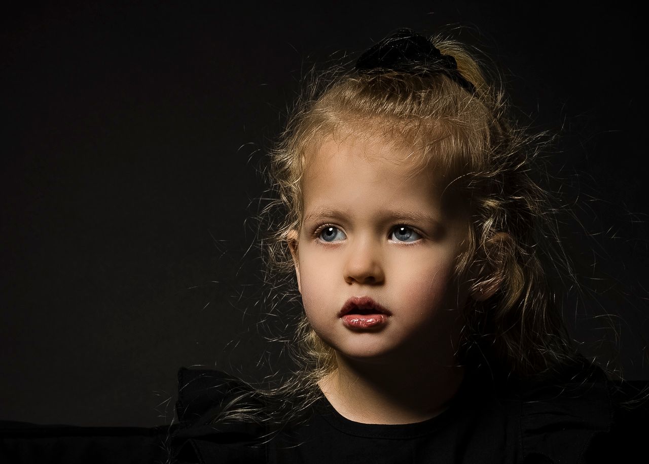 A child with curly blonde hair wearing a dark shirt poses against a black background in dramatic lighting.